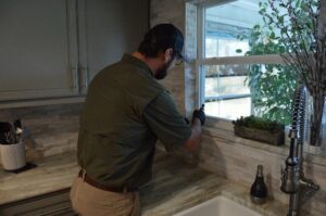 A Pro Care Lawn and Pest technician performing interior pest control treatment near a kitchen window in Jacksonville, FL.