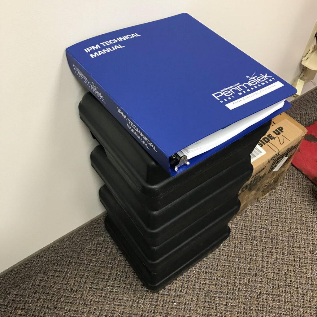 An IPM Technical Manual resting on a stack of black pest bait stations from Perimetek Pest Management in East Syracuse, NY.