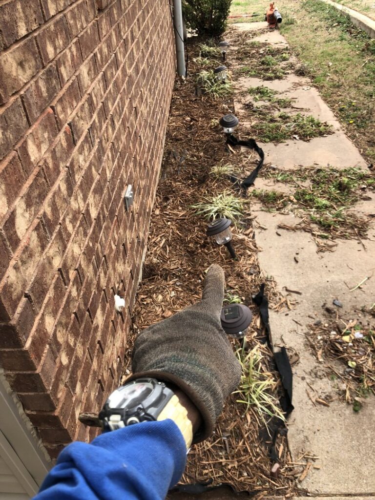 A gloved hand pointing at a freshly mulched landscape bed with new plants and solar lights by A & A Lawn Care Service LLC in Edmond, OK.