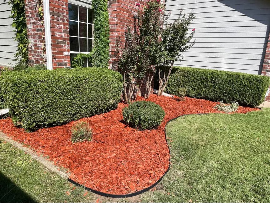 A neatly landscaped flower bed featuring vibrant red mulch and well-trimmed bushes, a service provided by Lande Lawn Care in Broken Arrow, OK.
