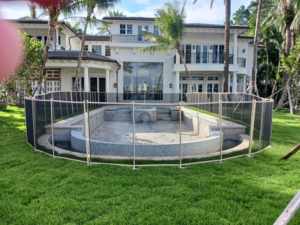 A large backyard pool secured with a light-colored mesh safety fence by All Brand Pool Fence in Fort Lauderdale, FL.