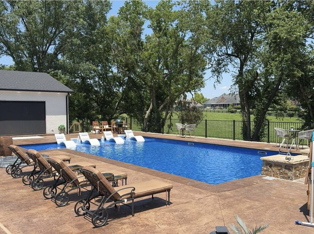 A large, custom-shaped inground pool with a wide concrete patio and lounge chairs, a design by Riverstone LLC in Bowling Green, KY.