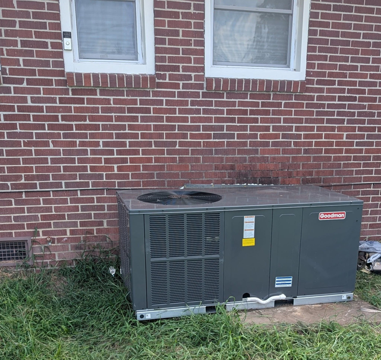 A large Goodman outdoor AC condenser unit installed next to a brick building by Prestige Heating Cooling & Appliances Llc in Sumter, SC.