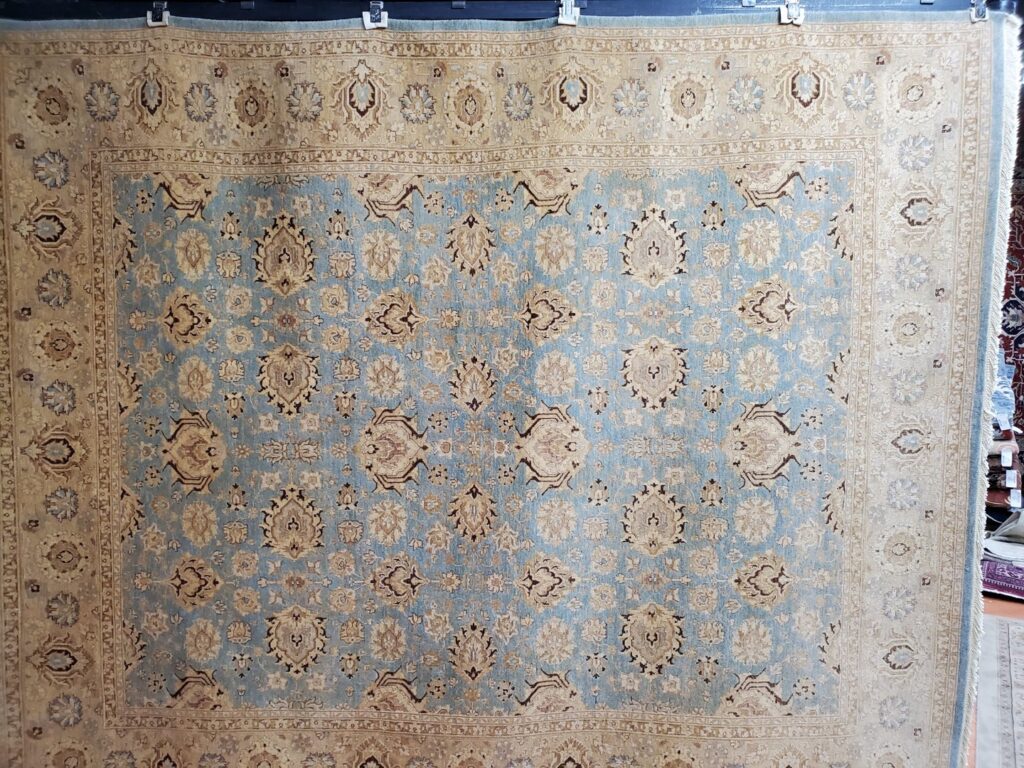 A large light blue and beige patterned rug hanging for display at Khazai Rugs in Lexington, KY