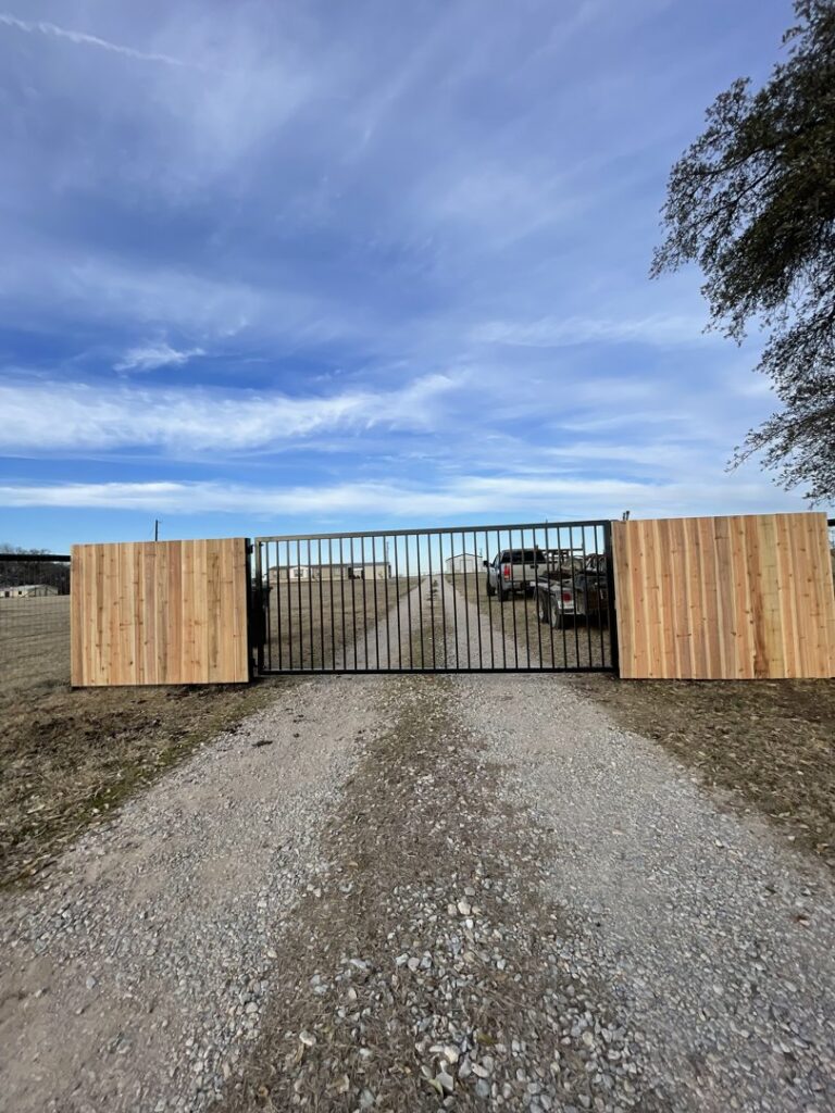 A large black metal gate with wood fence sections on a gravel driveway by Backyard Builders in Rockford, MI.