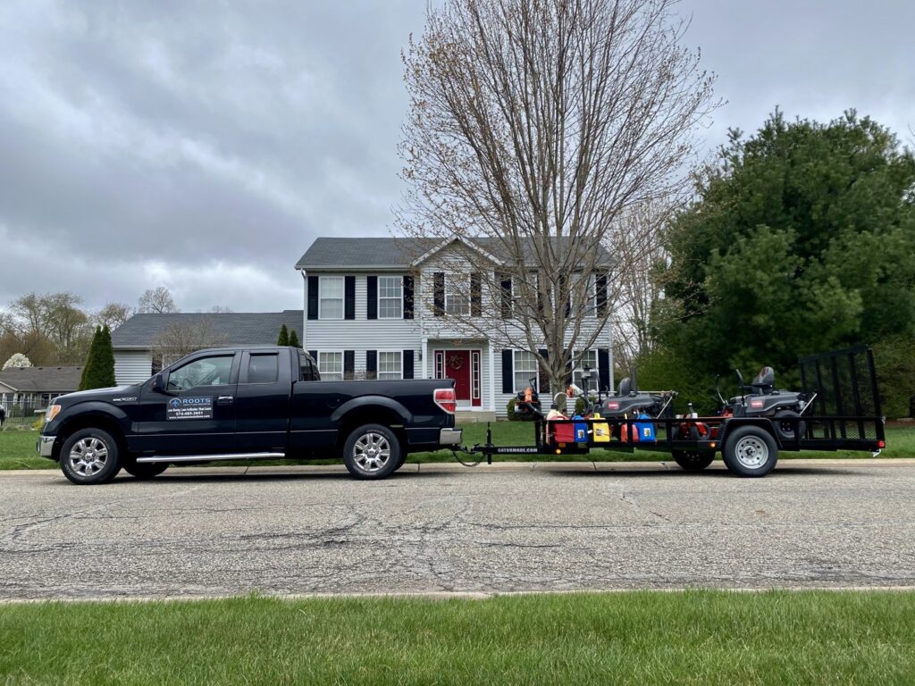 A Roots Lawn Care Solutions truck and trailer with professional equipment parked on a residential street in Granger, IN.