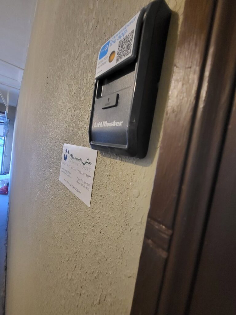 A LiftMaster wall control panel with a Mountain Fox Garage Doors business card, showing a completed installation in Colorado Springs, CO.
