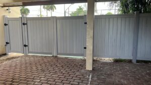 A newly installed light grey vinyl fence with a gate under a patio by Palm's Fence of Miami, FL.