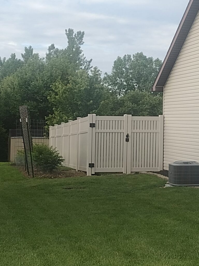 A light-colored vinyl privacy fence with a gate installed next to a house by Nailed It Fencing and Exteriors in Evansville, IN.