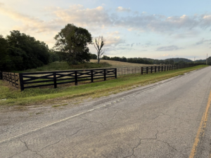 A long black wooden fence with a metal gate installed alongside a rural road by The Fence Painter in Lynnville, TN.