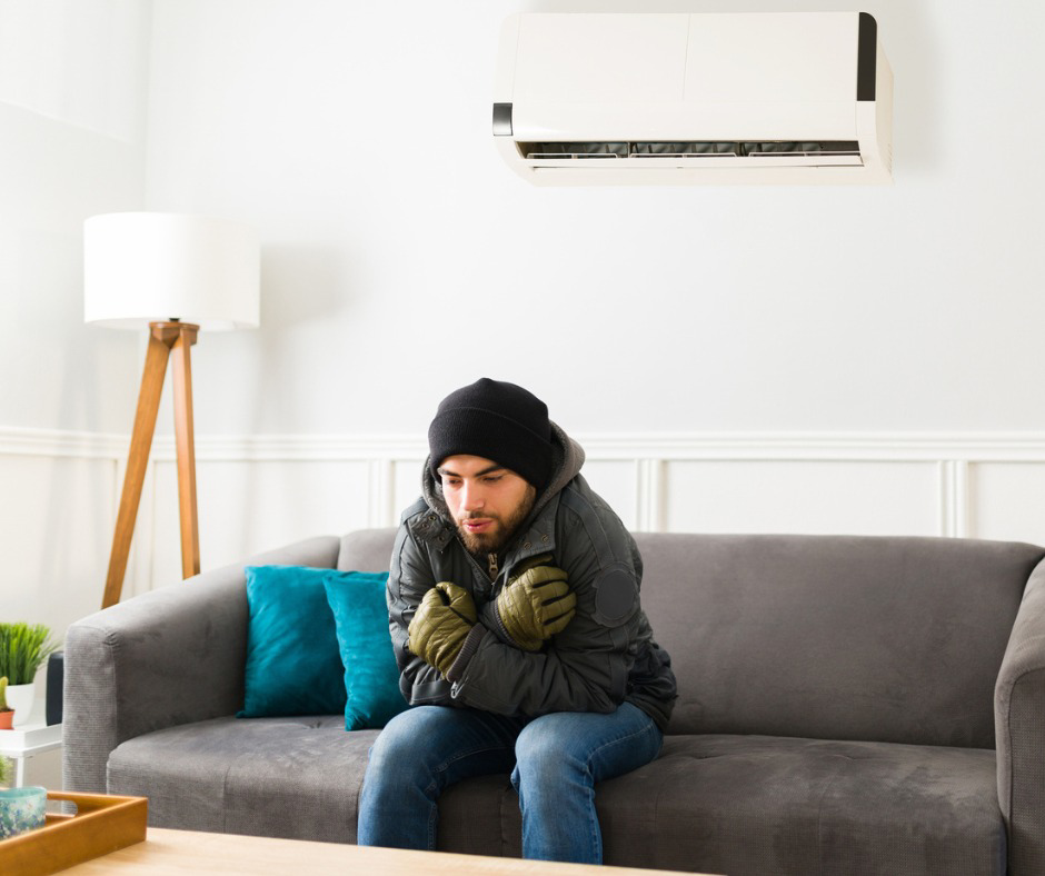 A man shivering in a cold room with a mini-split AC unit on the wall, illustrating a common heating problem Ed's Comfort Solutions, Inc solves in Callahan, FL.