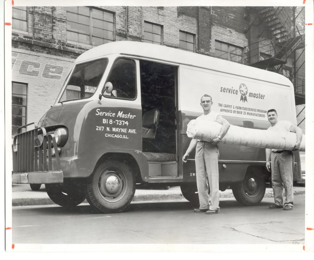 Two men carrying a rolled carpet next to a vintage Service Master van, showcasing carpet services by ServiceMaster Albino in Waterbury, CT.