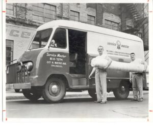 Two men carrying a rolled carpet next to a vintage Service Master van, showcasing carpet services by ServiceMaster Albino in Waterbury, CT.