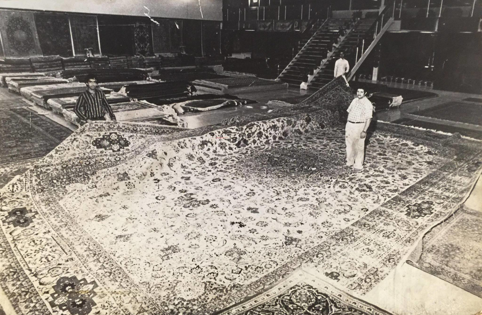 Two men displaying a large, intricately patterned Persian rug at Tousi Rugs in Los Angeles, CA.