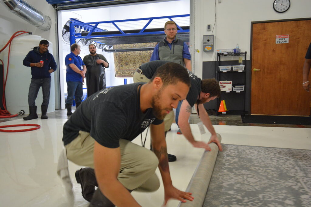 Men rolling up a large area rug, preparing it for care or storage at the Association of Rug Care Specialists in Salem, OR