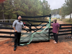Two men standing with a new black metal gate at a fence line, ready for installation by All With Us LLC in Green Cove Springs, FL.