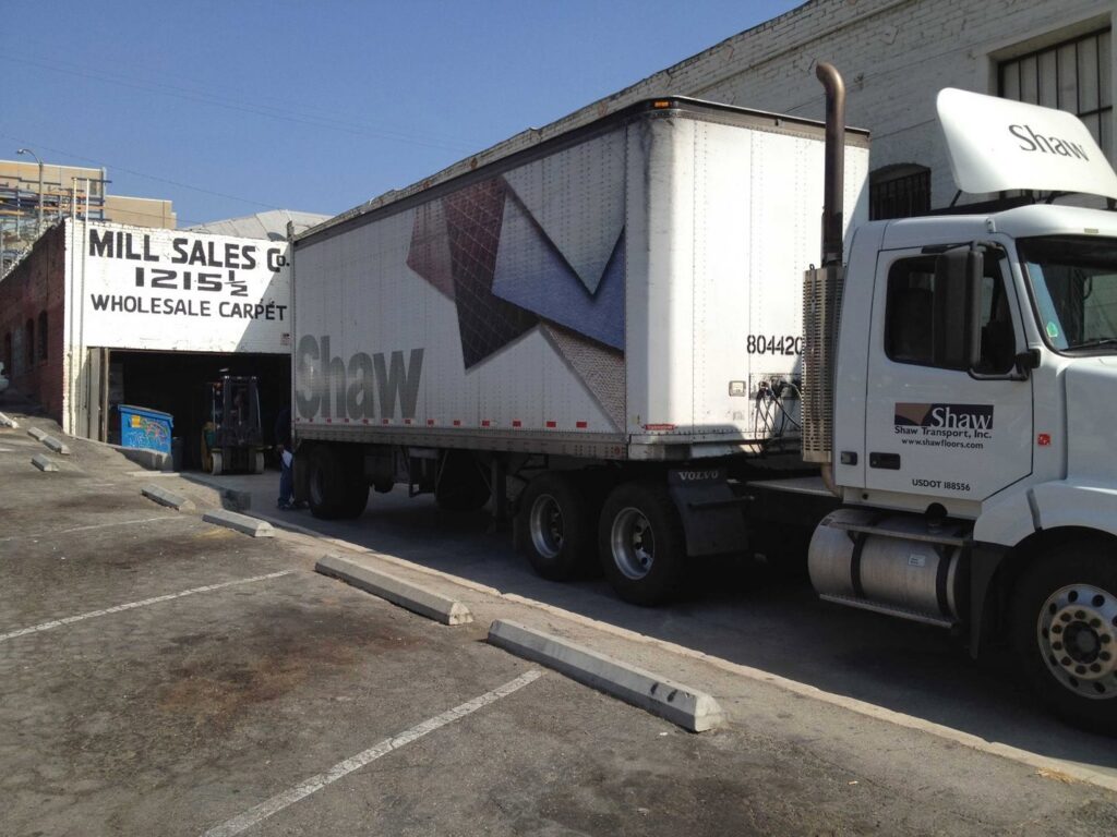 The exterior of Mill Sales Co. building with a sign reading "WHOLESALE CARPET" and a Shaw delivery truck in Los Angeles, CA