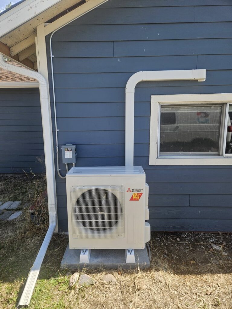 An outdoor mini-split condenser unit installed next to a blue house by 406 Heating & Cooling LLC in Missoula, MT.