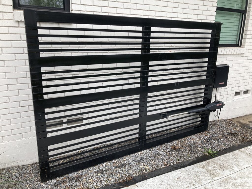 A modern automatic black metal sliding gate installed against a white brick wall by Craftsman Automatic Gates in Dallas, TX.
