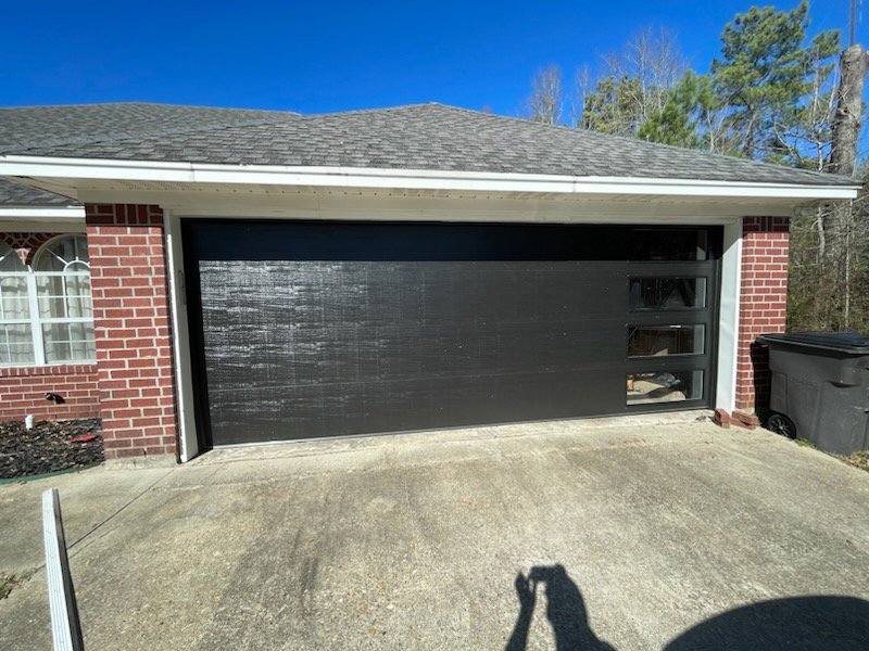 A modern black garage door with horizontal panels and windows installed on a brick home by Geaux Overhead Door in West Monroe, LA.