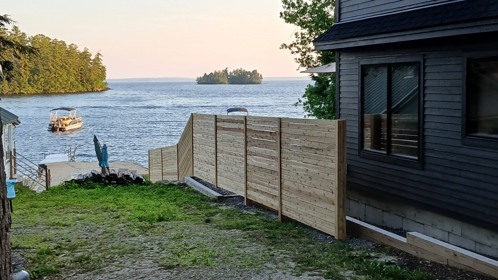 A modern horizontal slat wooden fence installed next to a lakeside home by Creative Fence in Casco, ME.