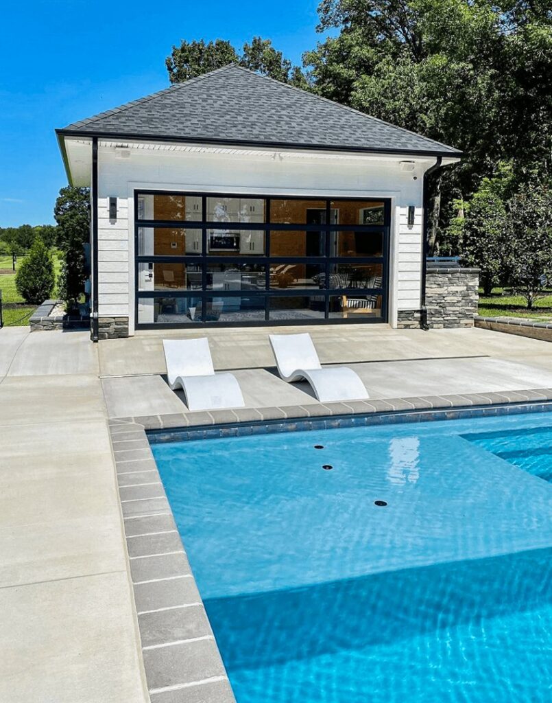 A modern pool house featuring a full-view glass garage door overlooking a swimming pool, installed by Monroe Overhead Doors in Monroe, LA.