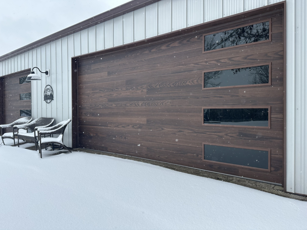 A modern wood-look garage door with windows, installed by American Eagle Garage Doors in Greeley, CO, covered in light snow.