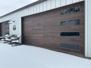 A modern wood-look garage door with windows, installed by American Eagle Garage Doors in Greeley, CO, covered in light snow.