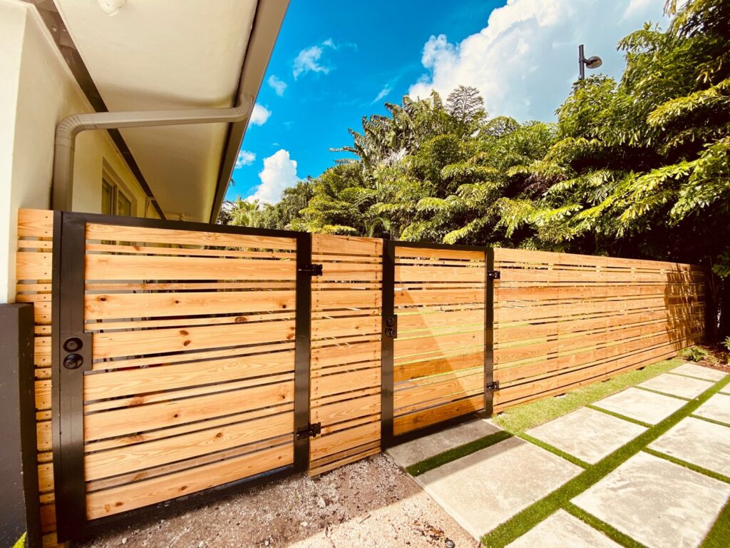 A modern wooden fence with black metal posts and a gate installed in a tropical setting by Miami Fence Installation Contractors in Miami, FL.