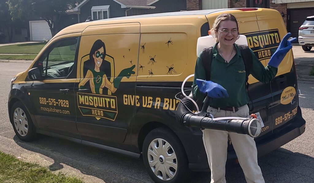 A smiling Mosquito Hero female technician with a backpack sprayer stands by a branded service van in Champaign, IL.