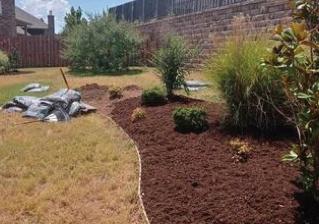 A newly mulched flower bed with bags of mulch nearby, showing landscaping work by From The Roots Lawn Service in Edmond, OK.