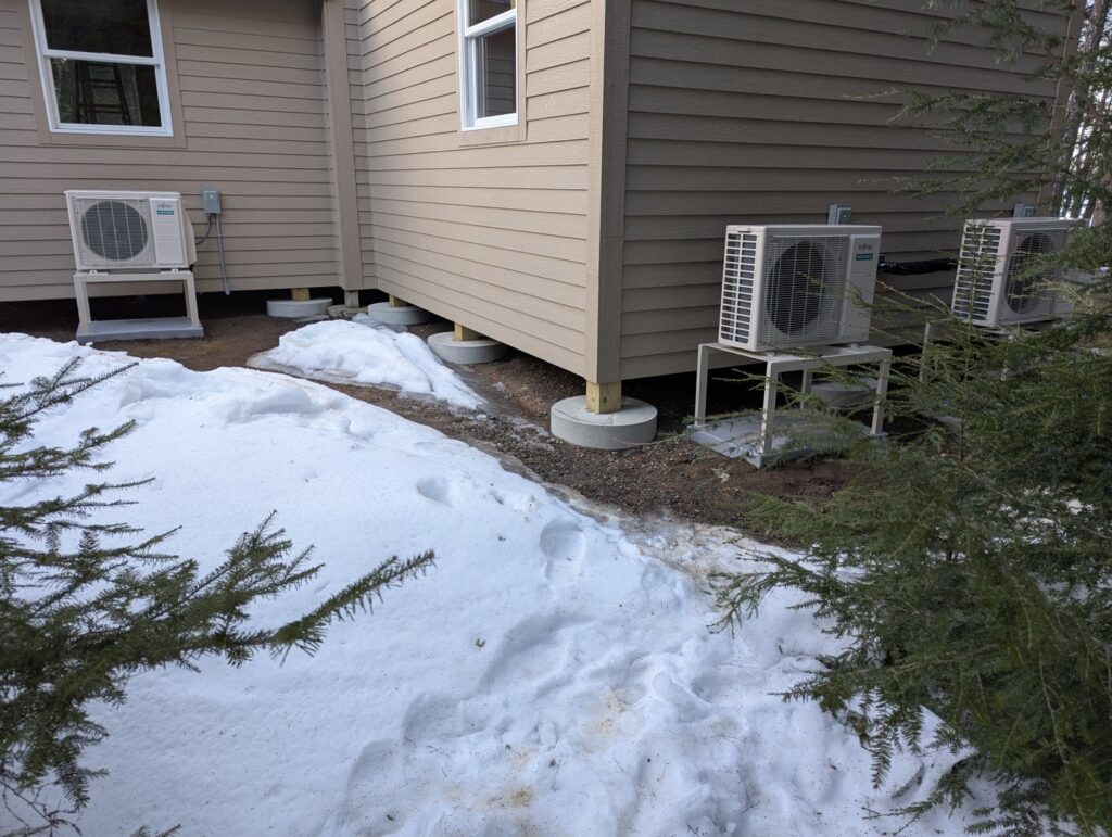 Multiple outdoor heat pump units installed on the side of a house in winter by Kinney Family Heat Pumps in Augusta, ME.