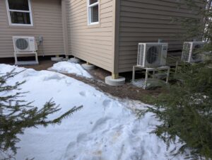 Multiple outdoor heat pump units installed on the side of a house in winter by Kinney Family Heat Pumps in Augusta, ME.