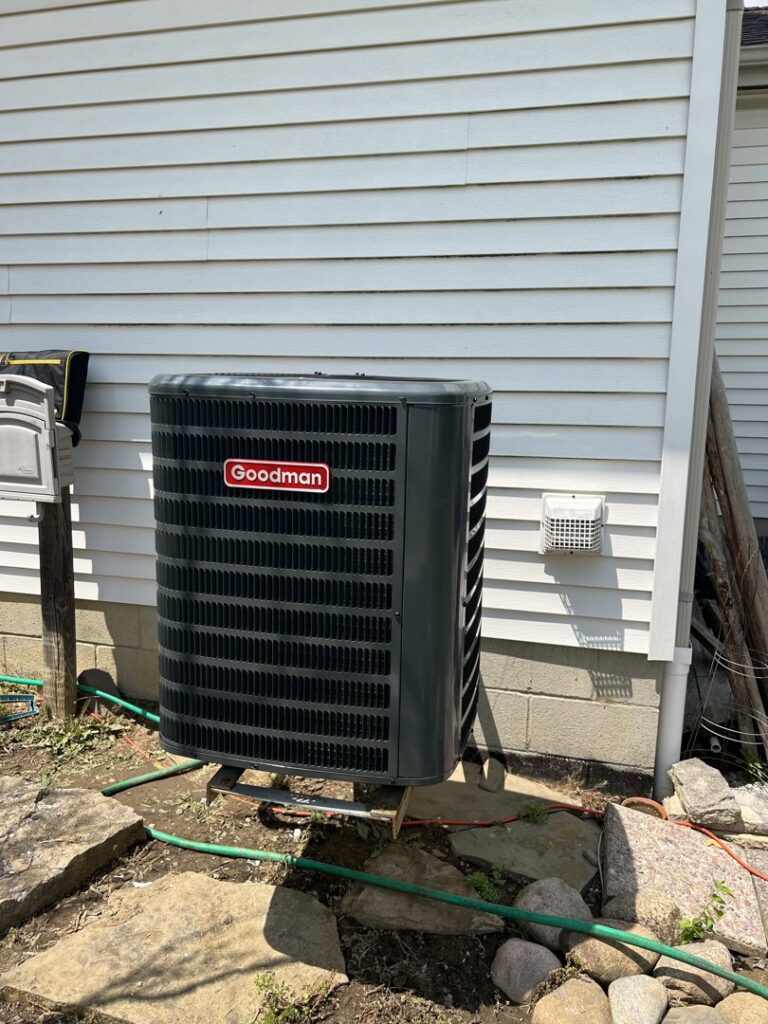 A new Goodman outdoor AC unit installed next to a house exterior with rocks and a garden hose by Buckeye Mechanical Construction in Columbus, OH.