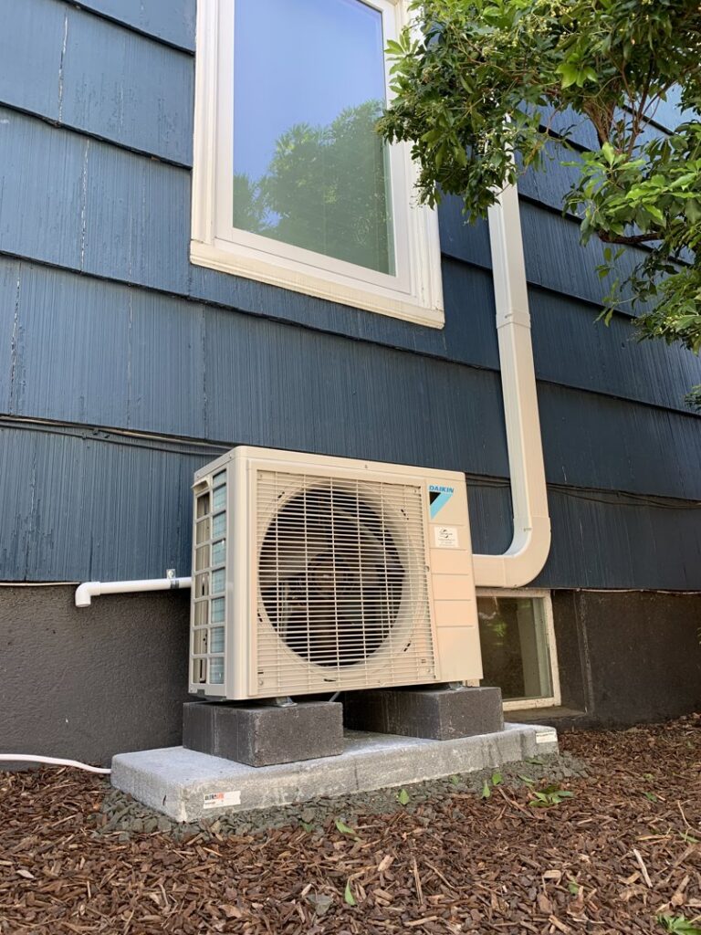 A new outdoor heat pump unit installed on a concrete pad next to a blue house by The Heat Pump Store in Portland, OR.