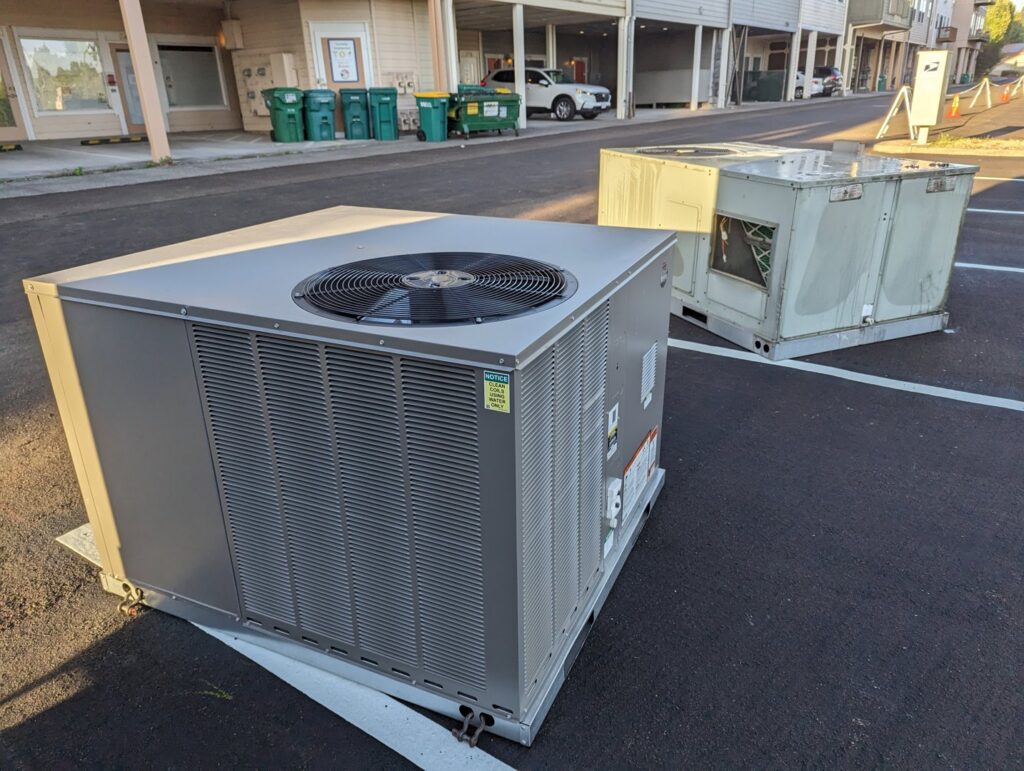 A new outdoor HVAC unit and an old rooftop unit in a parking lot, indicating a replacement job by Gresham Heating and Air Conditioning in Wood Village, OR.