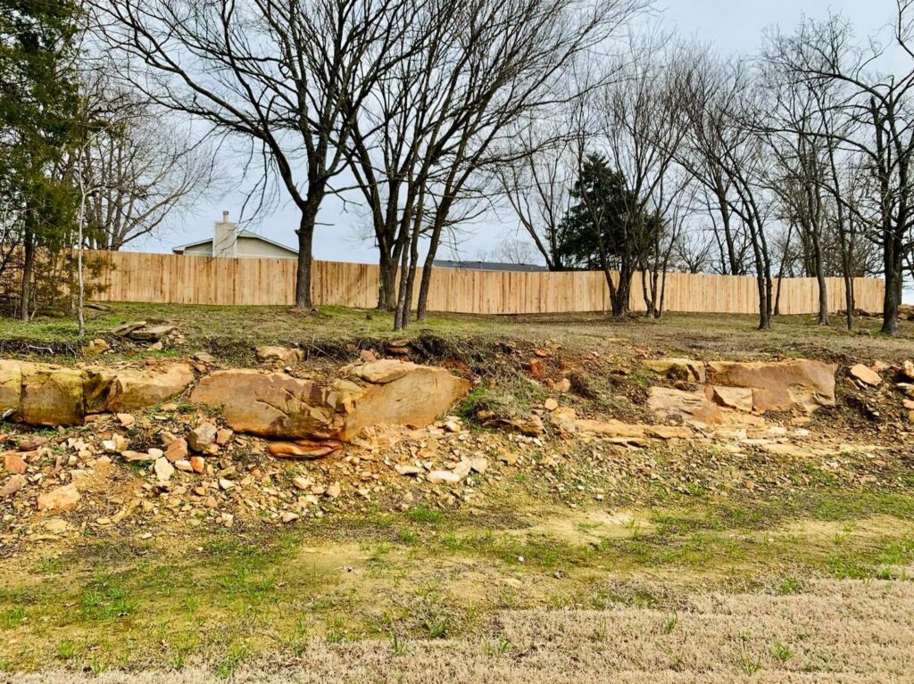 A newly installed wooden fence on a challenging rocky and sloped terrain by Ninety-One Services in Tulsa, OK