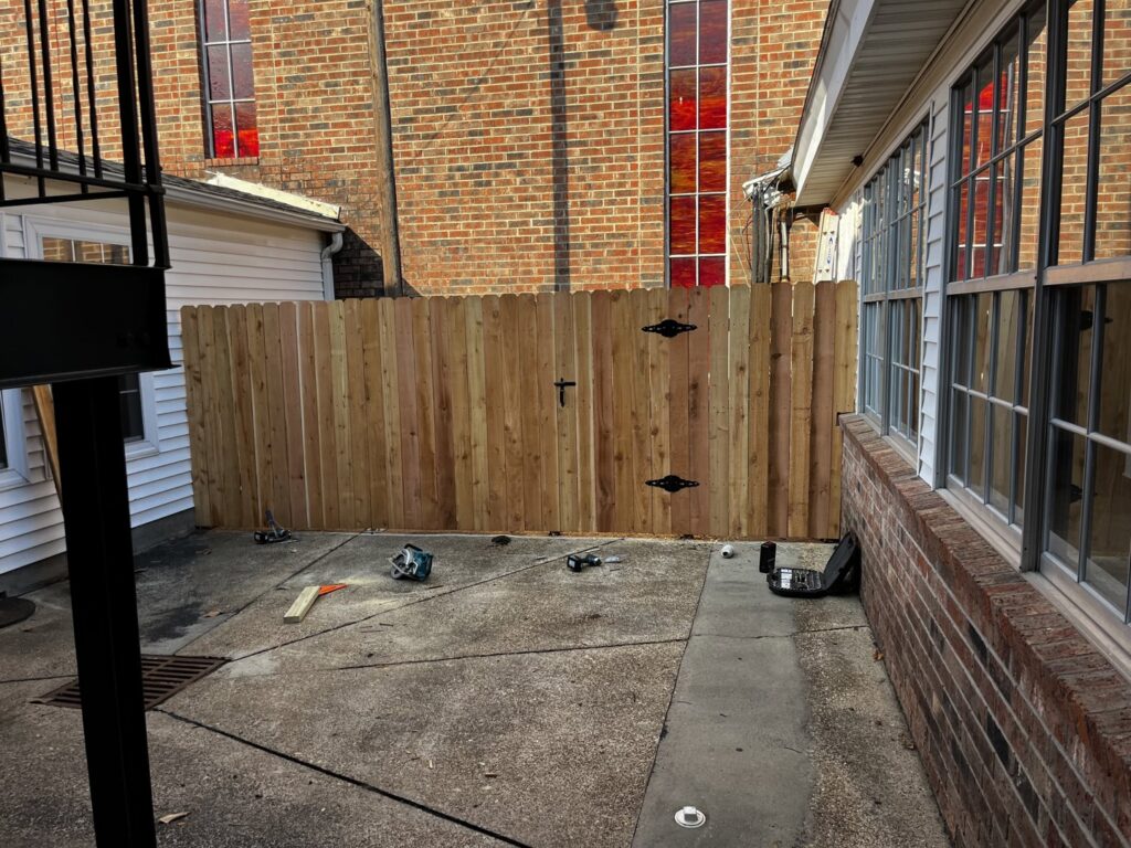A newly installed wooden privacy fence with a gate next to a brick building by Dallas' Custom Carpentry in Alexandria, LA.
