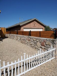 A newly installed wood fence on top of a stone retaining wall, with a white picket fence in the foreground by MJ Custom Fencing in Las Cruces, NM.