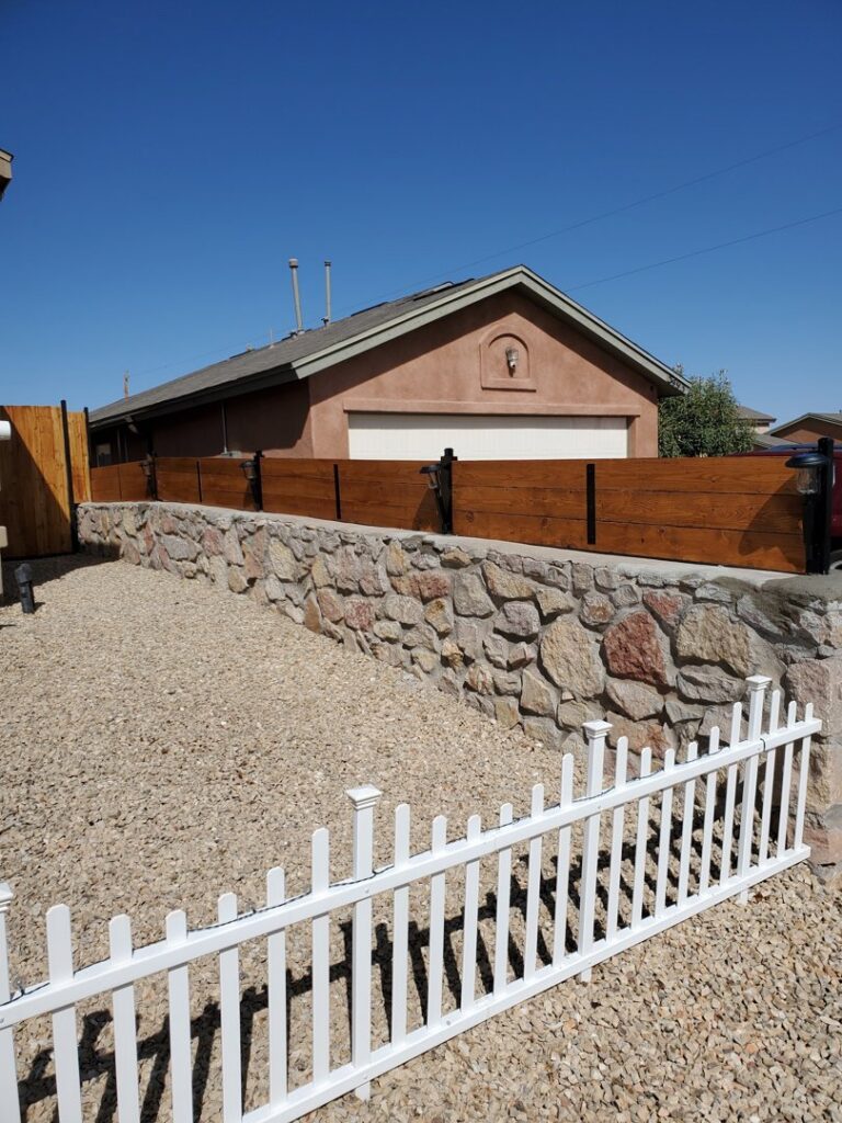 A newly installed wood fence on top of a stone retaining wall, with a white picket fence in the foreground by MJ Custom Fencing in Las Cruces, NM.