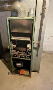 An old furnace unit with its cover removed, showing internal components and ductwork, before replacement by Cousins Heating & Cooling in Lansing, MI.