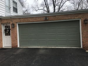 An older olive-green garage door on a brick residential house, showing potential for replacement by Geiszler Garage Door in Milwaukee, WI.