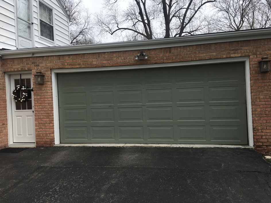 An older olive-green garage door on a brick residential house, showing potential for replacement by Geiszler Garage Door in Milwaukee, WI.