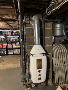 An old water heater or boiler unit with a flue pipe in a basement by Hampden County HVAC Inc. in Indian Orchard, MA