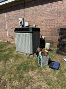 An older outdoor AC unit next to a brick wall with tools and a box, indicating service by Fort Worth Air Conditioning Co. Inc. in Fort Worth, TX.
