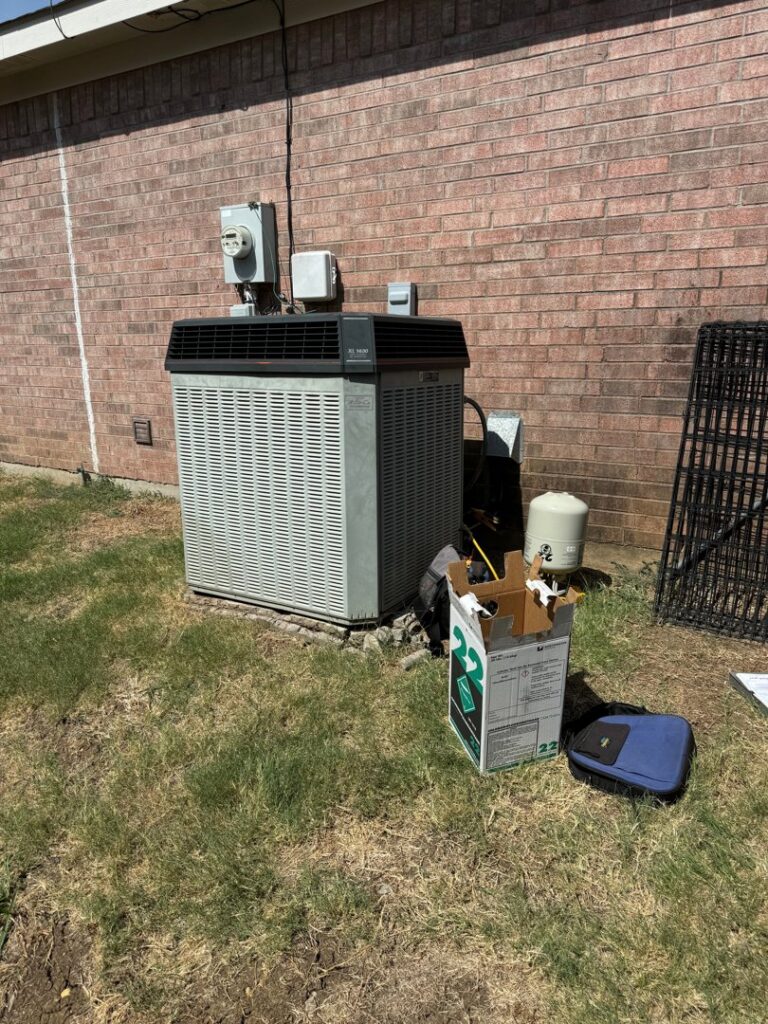An older outdoor AC unit next to a brick wall with tools and a box, indicating service by Fort Worth Air Conditioning Co. Inc. in Fort Worth, TX.