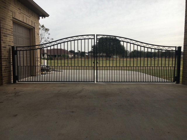 A large ornamental iron double gate installed at a driveway entrance next to a brick building by Texas Fence and Iron Co. in Houston, TX.