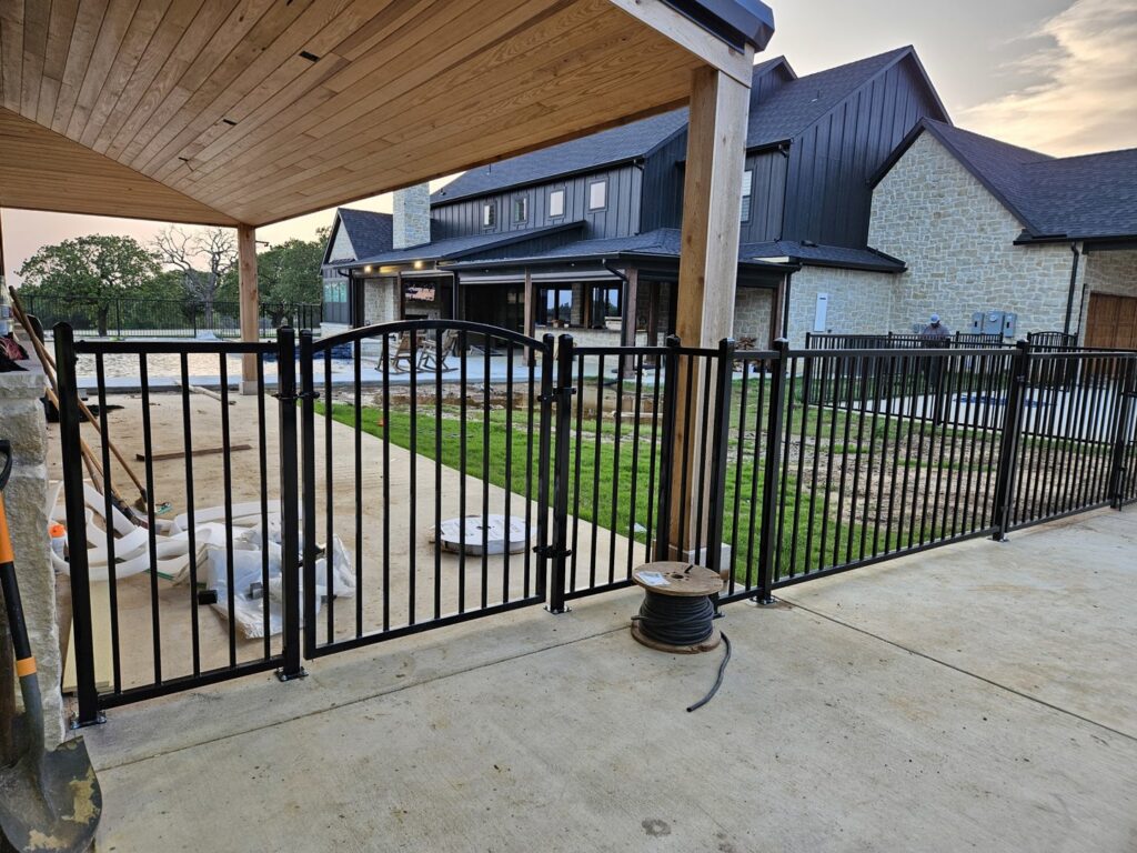 Black ornamental metal fence and gate enclosing a patio area next to a modern home, installed by QD & E Fencing in Fort Worth, TX.