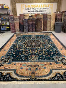 An ornate area rug displayed in the showroom of Mussallem Area Rug Specialist in Jacksonville, FL, with many rolled rugs.