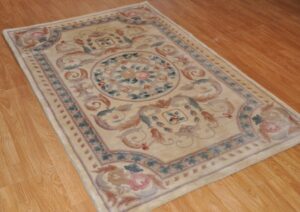 An elegant light-colored rug with an ornate floral and scroll pattern displayed on a wooden floor, a typical item for N J Green Rug Cleaning in Jersey City, NJ.
